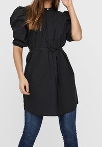 Schwarzes, übergroßes Hemdkleid aus Baumwolle mit Puffärmel, Knopfverschluss vorne und Taillengürtel, kombiniert mit schmalen Jeans.