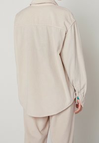 Light beige corduroy shirt with a collared neckline, long sleeves, and a curved hem. Textured fabric with vertical ribs and buttoned cuffs.