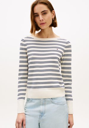 Kvinde iført en hvid langærmet sweater med tynde mørkeblå vandrette striber, kombineret med lyseblå jeans, stående foran en ensfarvet baggrund.