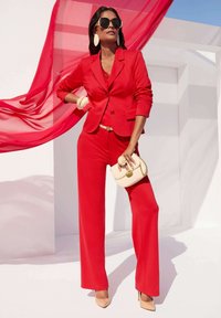 Roter maßgeschneiderter Anzug mit langen, weiten Hosen und taillierter Blazer. Glattes Material, kombiniert mit hautfarbenen Absätzen und einer cremefarbenen Handtasche. Auffällige Ohrringe.