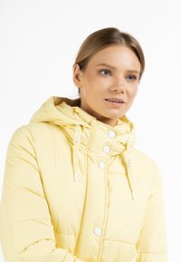 Gul pufferjakke med hætte, der har kviltet tekstur, for- og bagknapper, samt snorede detaljer. Glat, letvægtsmateriale.
