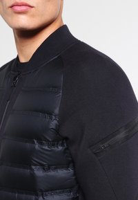 Černá bomber bunda s předním zapínáním na zip, polstrovanými částmi na trupu a hladkými, texturovanými pletenými rukávy s kapsou na zip na paži.