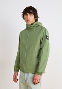 Green lightweight jacket with a front zipper, elastic cuffs, and adjustable hood. Features a rectangular patch on the left sleeve.
