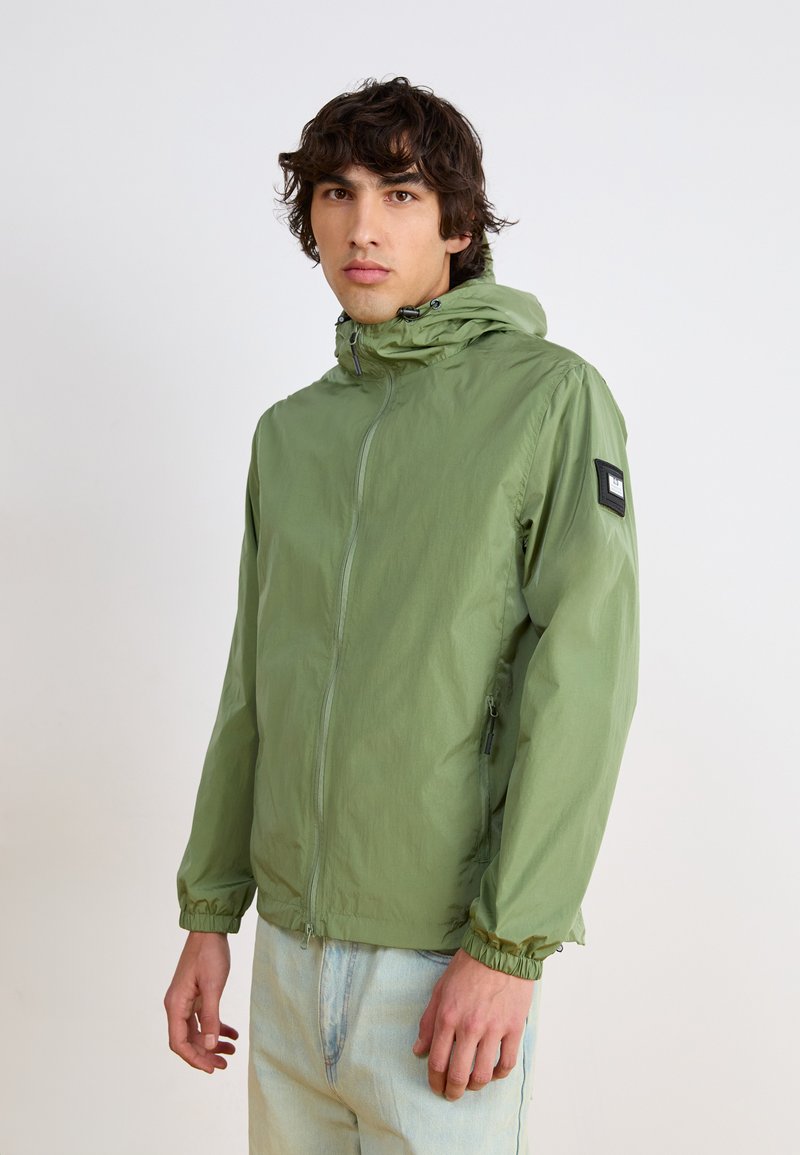 Green lightweight jacket with a front zipper, elastic cuffs, and adjustable hood. Features a rectangular patch on the left sleeve.