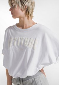 Camiseta blanca de manga corta y corte oversize hecha de algodón. Presenta un estampado texturizado con la palabra "ACTITUD" y un escote redondo, combinada con pantalones grises.