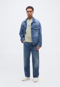 Άνδρας που φοράει ριγέ μπλε τζιν μπουφάν, ανοιχτό πράσινο πουκάμισο, μπλε τζιν και λευκά αθλητικά παπούτσια, στέκεται με το ένα χέρι στην τσέπη.