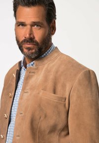 Giacca in suede marrone con colletto alto, caratterizzata da bottoni dorati e una piccola tasca sul lato sinistro. Indossata sopra una camicia a quadri blu.