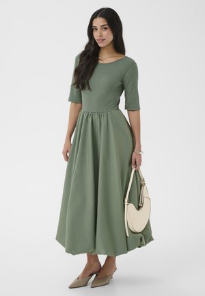 Femme portant une robe midi verte à manches mi-longues, des talons pointus beiges et un sac à main crème, posant devant un fond blanc uni.