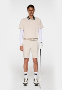 Beige pikétröja med kort ärm och randig krage, ljusbeiga shorts, vit långärmad undertröja och svart-vita golfsko, håller en golfklubba.