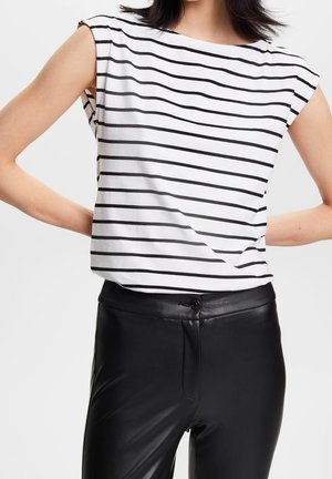 Kobieta ubrana w biały top bez rękawów z czarnymi poziomymi paskami i czarne skórzane spodnie z wysokim stanem, ramiona zgięte za plecami.