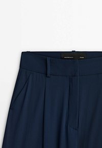 Pantalon bleu marine avec une ceinture plate, des poches latérales et un tissu lisse et texturé. L'étiquette indique « Massimo Dutti Studio ».
