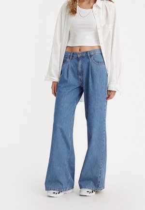 Kvinde iført lyseblå viddebukser, hvid crop top, åbnet hvid skjorte og hvide sneakers mod en ensfarvet baggrund.