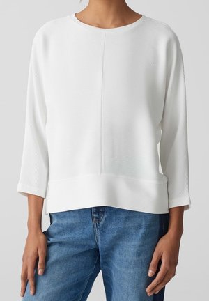 Vrouw draagt een wit geribd langarmpje met een centrale naad, gecombineerd met blauwe spijkerbroek, staand tegen een effen grijze achtergrond.