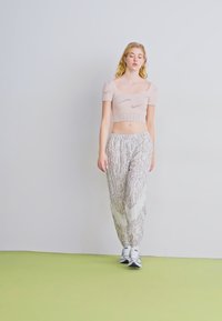 Gaiši rozā īsais tops ar īsām piedurknēm, apvienots ar brīviem, rakstainiem bēšiem biksēm. Modelis valkā tumšas sporta kurpes uz zaļa grīdas.