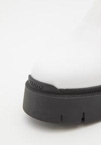 Botte en caoutchouc blanche à bout arrondi, dotée d'une semelle plateforme noire, d'accents texturés et du nom de la marque "HUGO" embossé sur le côté.
