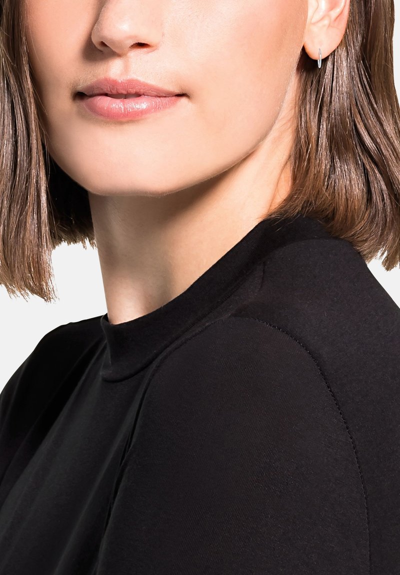 Top nero a collo alto con una texture liscia, caratterizzato da spalle aderenti e una silhouette pulita. Capelli corti e lisci e piccolo orecchino a cerchio in argento visibile.