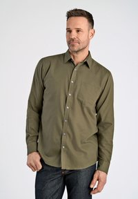 Camisa de manga larga verde oliva con un bolsillo frontal, cuello abotonado y botones plateados, combinada con jeans azul oscuro.