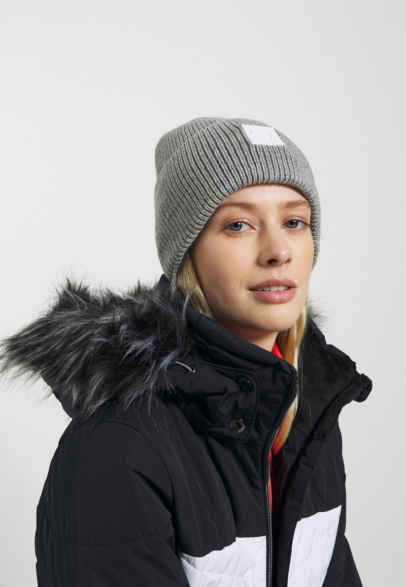 Grå stickad mössa med ribbat mönster, prydd med en vit logotyp. Bärs tillsammans med en svart jacka med pälsfodrad huva.