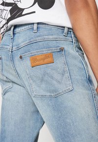 Svijetloplave traperice s izblijedjelim izgledom, koje imaju smeđi kožni oznaku s "Wrangler" logotipom i prepoznatljiv obrazac šivanja na stražnjem džepu.