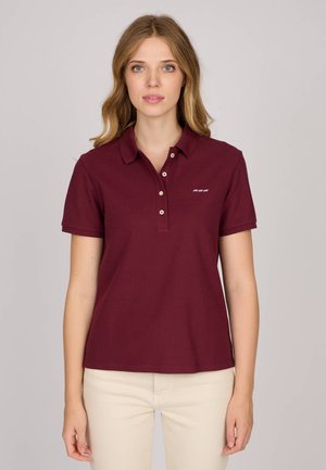 Femme aux cheveux blonds portant un polo à manches courtes bordeaux et un pantalon beige clair, se tenant devant un fond uni clair.