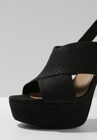 Sandale à plateforme compensée noire avec des brides croisées texturées et un bout ouvert, présentée sur un fond gris clair uni.