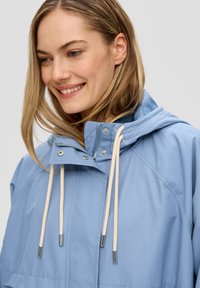 Veste imperméable bleu clair avec capuche, dotée d'une fermeture éclair à l'avant, de cordons de serrage et d'accents métalliques. Texture lisse et design moderne.