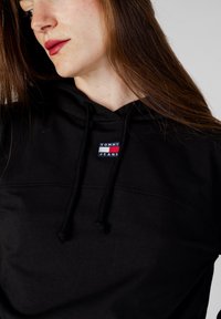 Felpa nera in cotone con tasca frontale, cappuccio con coulisse e un'etichetta logata rettangolare con la scritta "TOMMY JEANS" in blu e rosso.