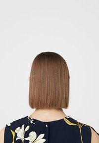 Cheveux bruns et raides coupés en carré droit. Le sujet porte un haut bleu marine foncé à motifs floraux. L'arrière-plan est blanc uni.