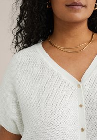 Cardigan blanc en tricot avec un décolleté en V, des boutons et des manches courtes. Le tissu présente un motif texturé.