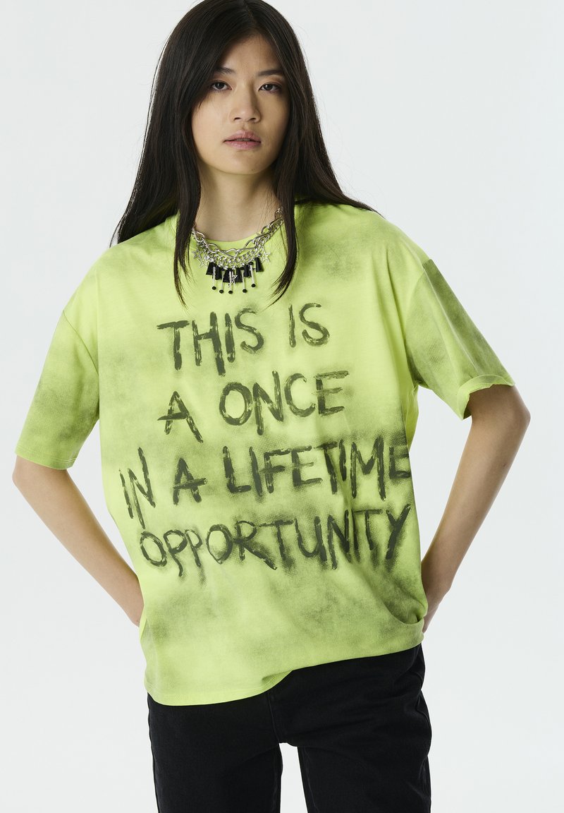 Camiseta de gran tamaño en verde neón con un texto pintado con spray en negro que dice "ESTA ES UNA OPORTUNIDAD ÚNICA EN LA VIDA". Mangas cortas.