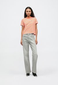 Woman standing wearing orange and white striped t-shirt, gray wide-leg jeans, and black heeled boots against white background.