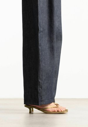 Personne portant un jean droit en denim foncé et des sandales à talon kitten vert olive avec fines brides, debout sur un sol lisse.