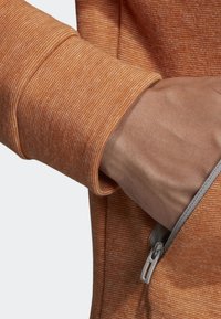 Sweat-shirt en tissu orange texturé avec une poche zippée et des poignets côtelés, montrant un gros plan de la main qui atteint la poche.