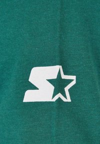 A zöld kötött anyag középpontjában egy nagy fehér "S" betű és egy csillag található, kiemelve merész dizájnját és textúráját.
