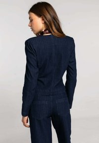 Blazer de mezclilla azul marino con una silueta ajustada, sutiles rayas verticales, hombreras estructuradas y bolsillos traseros en pantalones a juego.
