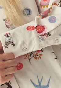 Valge kangast särk, millel on värvikad tikitud mustrid, sealhulgas karud, ringid ja lillemotiivid. Mansetil nupu detail.