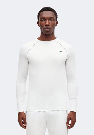 Hombre con una camisa blanca ajustada de manga larga con un pequeño logo verde de cocodrilo en el pecho izquierdo, de pie frente a un fondo liso.