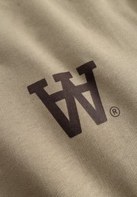 Beige stof med et mørkebrunt trykt logo "W" med et registreret varemærkesymbol. Materialet har en glat, struktureret overflade.