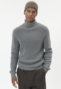Jauns vīrietis, tērpies pelēkā ribotā turtlenecka džemperī, brūnās biksēs un bēšā adītā cepurē, stāv uz neitrālas baltas fona.