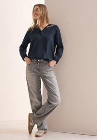 Marineblaue, strukturierte Bluse mit langen Ärmeln, kombiniert mit grauen, weit geschnittenen Jeans und grauen Sneakers mit beigen Akzenten; stehend gegen eine Wand.