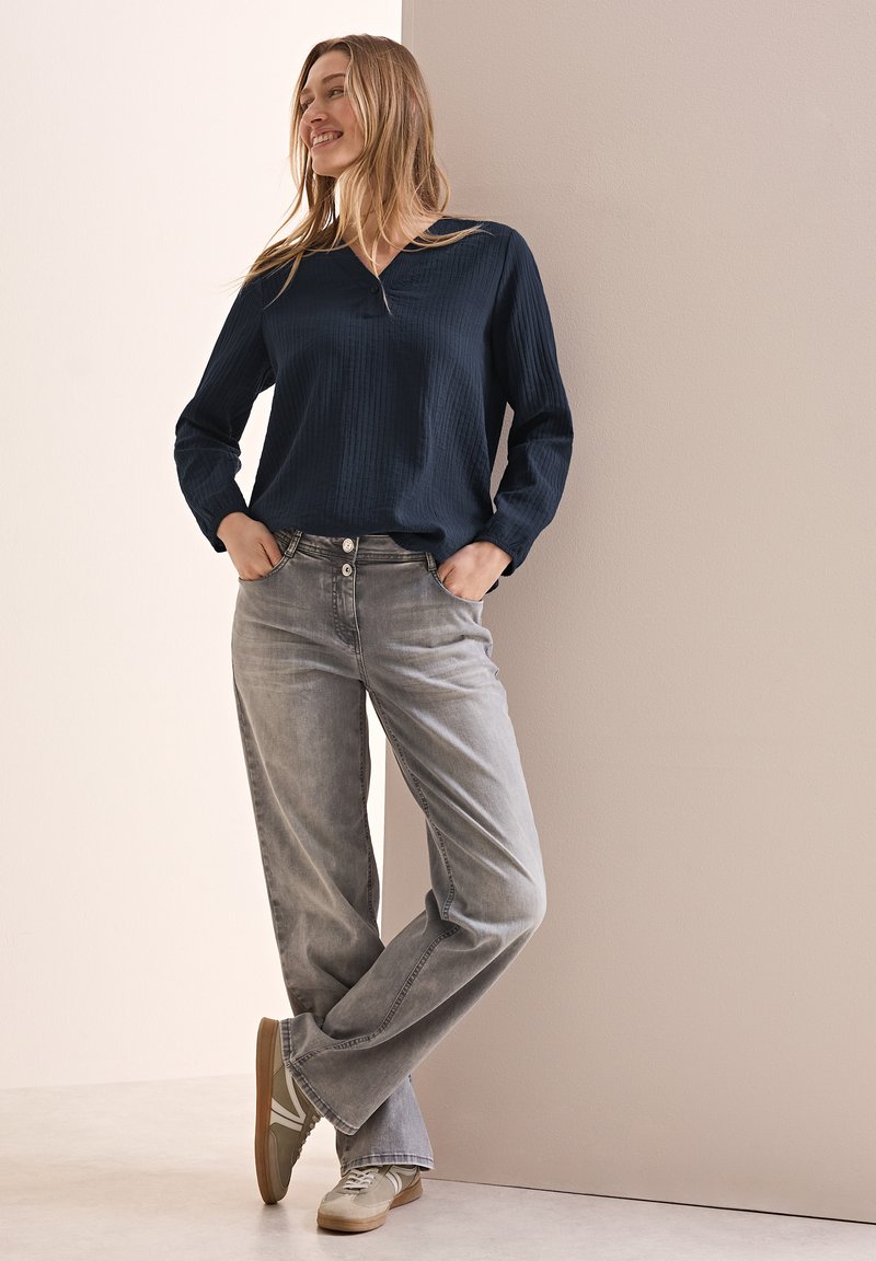Marineblaue, strukturierte Bluse mit langen Ärmeln, kombiniert mit grauen, weit geschnittenen Jeans und grauen Sneakers mit beigen Akzenten; stehend gegen eine Wand.