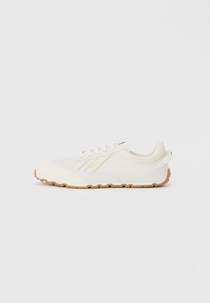 Sneakers athletic bianchi con una finitura liscia e texture, dotati di suola in gomma e un sottile logo dorato sul lato. Design minimalista.