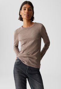 Donna che indossa una maglietta taupe a maniche lunghe e jeans neri, in piedi con le mani in tasca su uno sfondo chiaro e semplice.