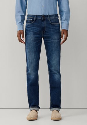 Man die een slim fit donkerblauwe spijkerbroek draagt, opgerold bij de enkels, een lichtblauw overhemd met knopen en beige instapschoenen.