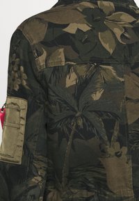Kombinovaná bunda s tmavě zelenými a hnědými květinovými vzory, náprsní kapsou na levém rukávu a texturovaným povrchem látky.