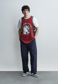 Ung mann med krøllete hår iført en rød basketballtrøye over en hvit t-skjorte, mørke cargobukser og svarte og hvite joggesko, stående med hendene i lommene.