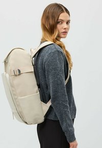 Beige ryggsäck med vadderade remmar, slät yta och sidoficka. Bärs över en grå tröja, vilket visar en strömlinjeformad design.