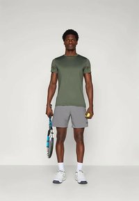 Camiseta deportiva verde oliva y pantalones cortos gris claro. Sosteniendo una raqueta de tenis y una pelota de tenis amarilla. Zapatillas blancas con acentos azules.