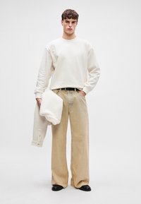 Λευκό πουλόβερ με χαμηλούς ώμους, μπεζ παντελόνι φαρδύ με διακριτική ραφή μπροστά και μαύρα παπούτσια. Κρατάει ένα λευκό υφαντό εξωτερικό ύφασμα.