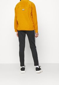 Gul fleece-tröja med en bakficka, ihop med svarta slim-fit byxor med vita sidostripe och svarta sportskor.